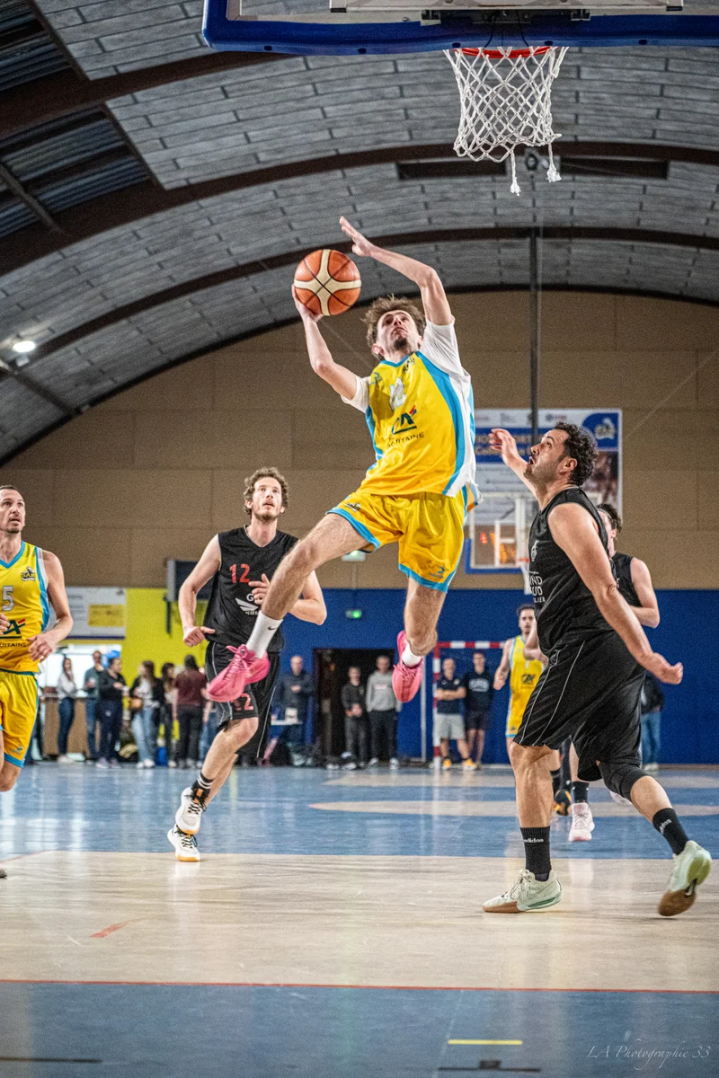 Joueur de basket en maillot jaune sautant pour un tir face à un défenseur en maillot noir lors d'un match en salle