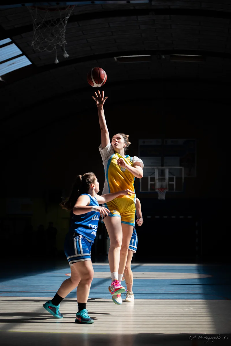 Joueuse de basket en maillot jaune et blanc effectuant un tir en suspension lors d'un match en salle