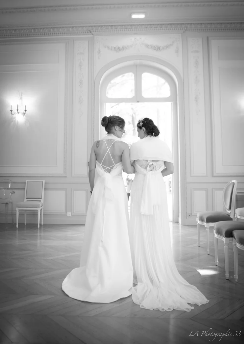 Photo en noir et blanc de deux mariées de dos, bras dessus bras dessous, dans un salon d'apparat