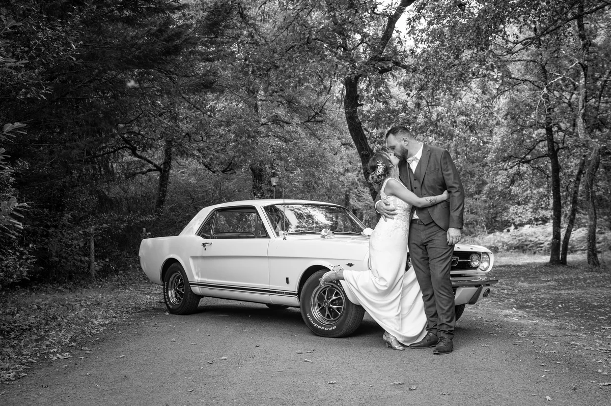 Photo en noir et blanc de mariés s'embrassant devant une voiture de collection