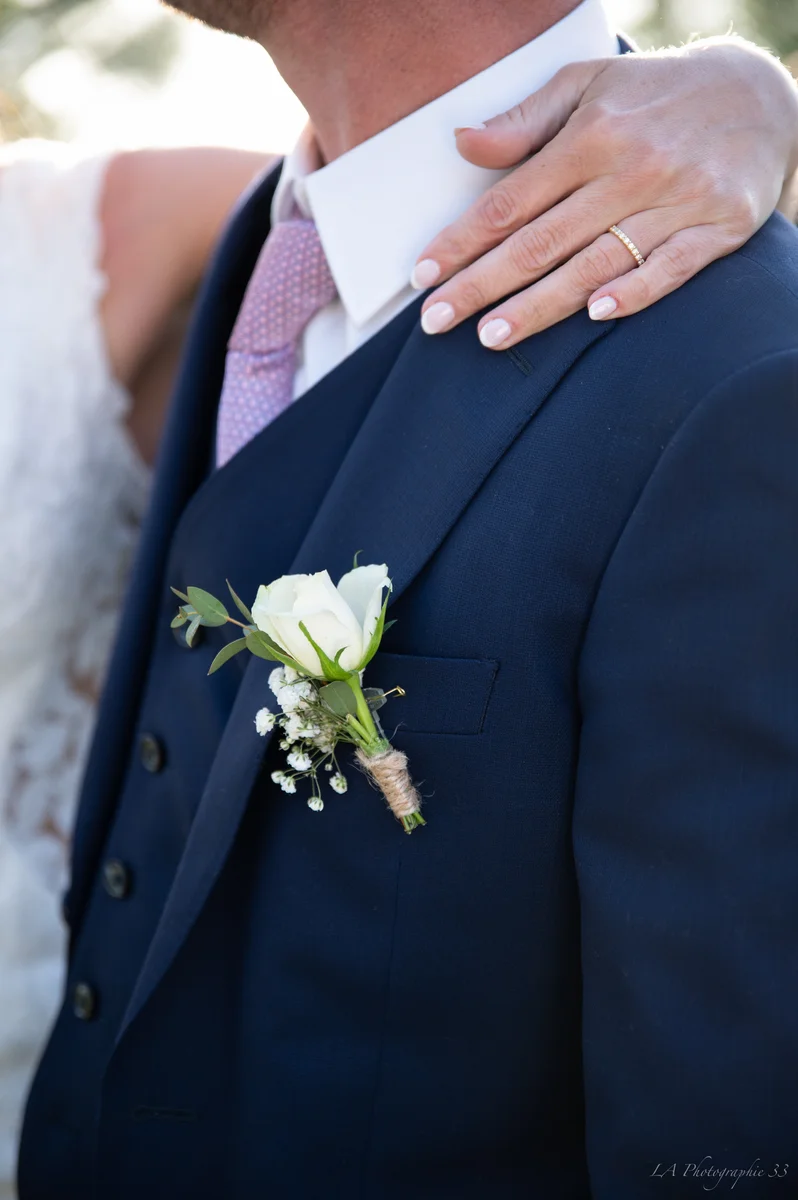 Gros plan sur une boutonnière de rose blanche et la main de la mariée sur l'épaule du marié