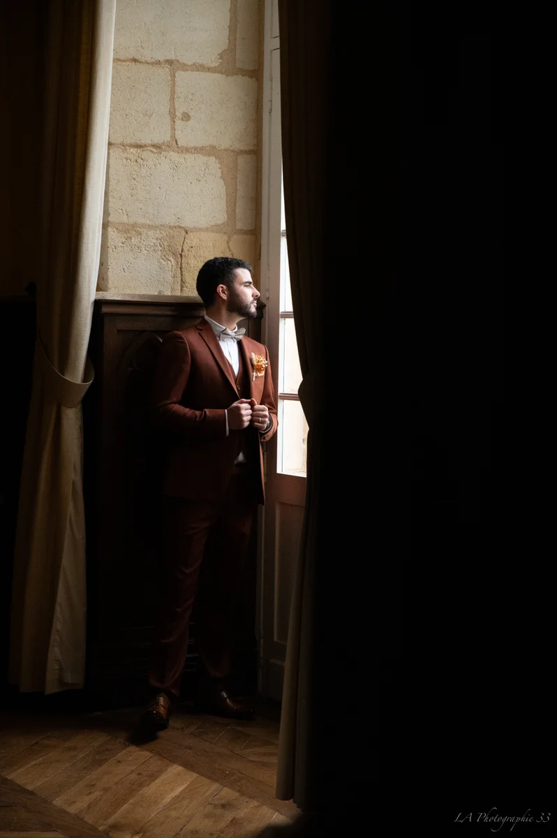 Marié en costume bordeaux regardant par une fenêtre, lumière douce