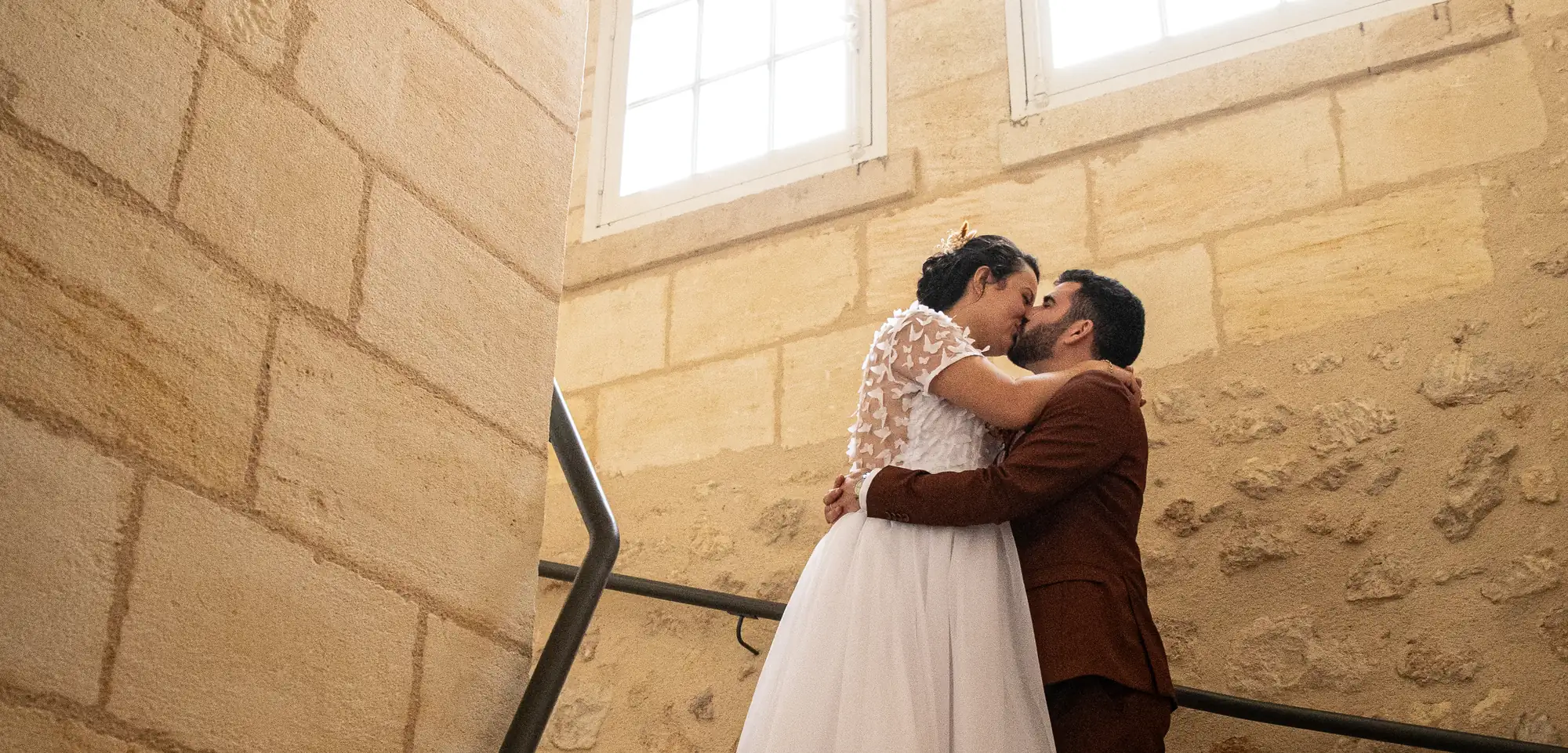 Mariés s'embrassant dans un escalier en pierre dorée, la mariée en robe blanche à appliqués papillons et le marié en costume bordeaux