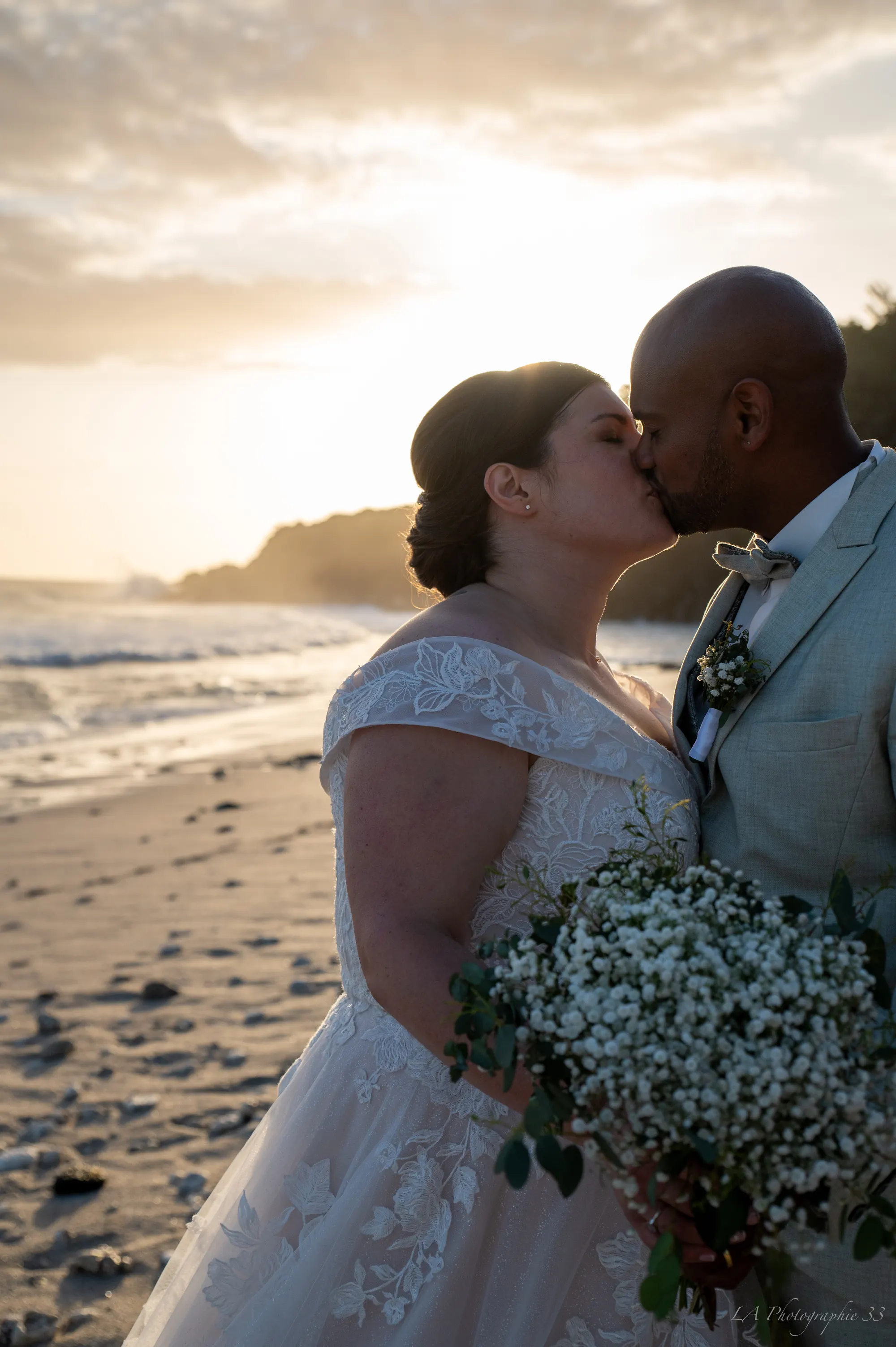 Mariés s'embrassant sur la plage au coucher de soleil, bouquet de fleurs blanches au premier plan