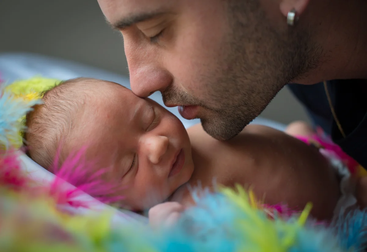 Papa déposant un baiser sur le front de son nouveau-né endormi sur un fond de plumes colorées