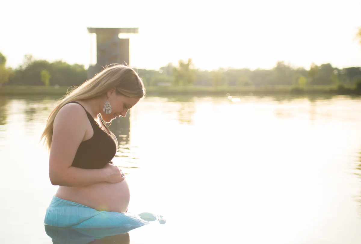 Femme enceinte de profil au bord d'un lac au coucher du soleil, regardant son ventre