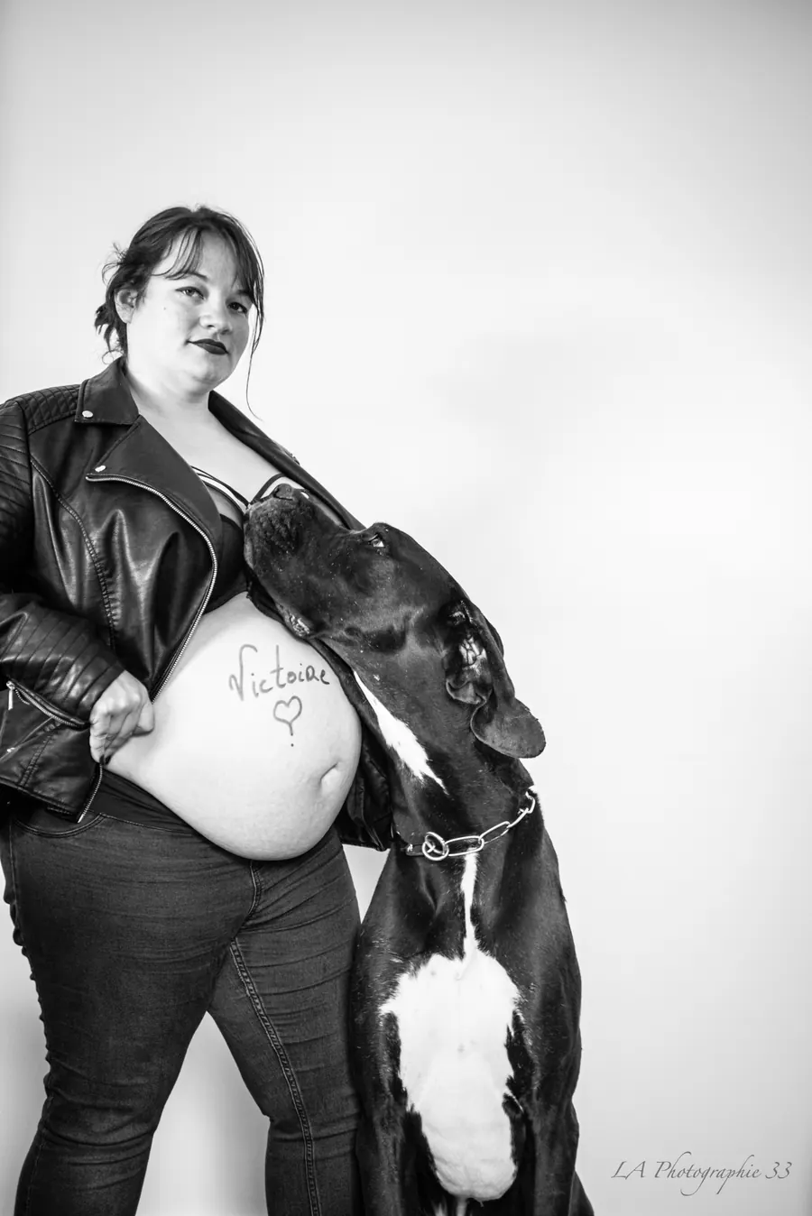Future maman en veste de cuir avec son chien, prénom du bébé écrit sur le ventre, photo en noir et blanc