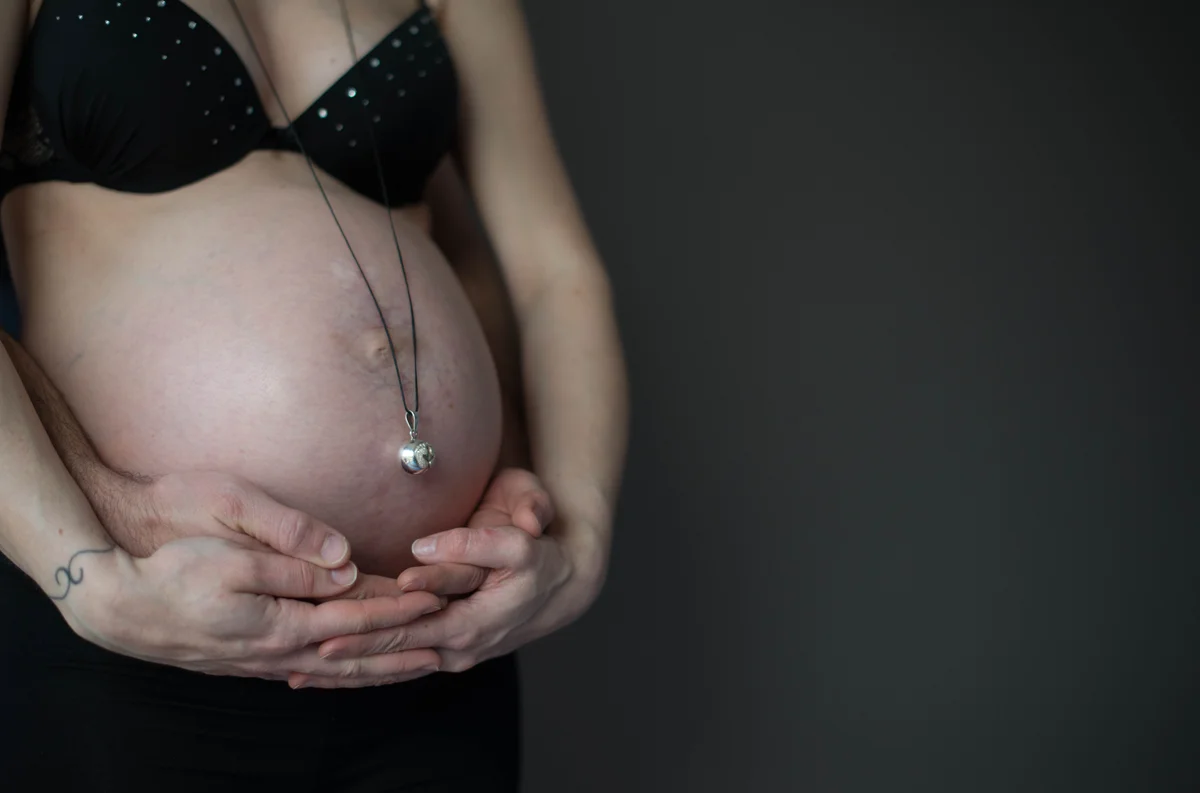 Ventre de femme enceinte encadré par ses mains, avec un collier pendentif cœur, fond sombre