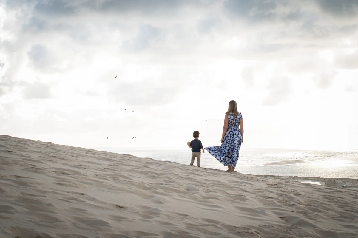 Mère et enfant de dos descendant une dune de sable vers l'océan, robe longue à fleurs bleues au vent