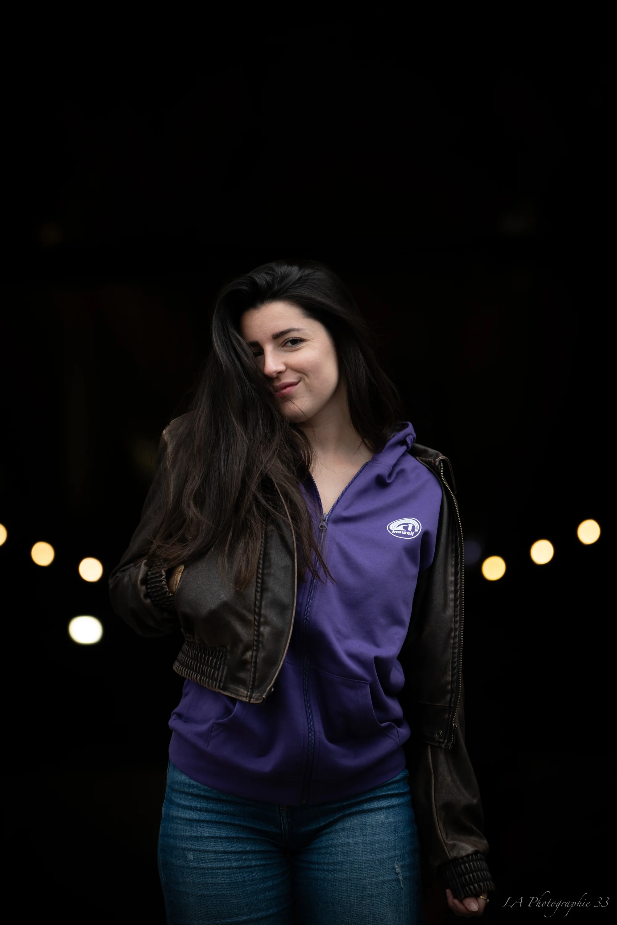 Jeune femme aux cheveux bruns souriante, portrait de nuit avec guirlande lumineuse en arrière-plan