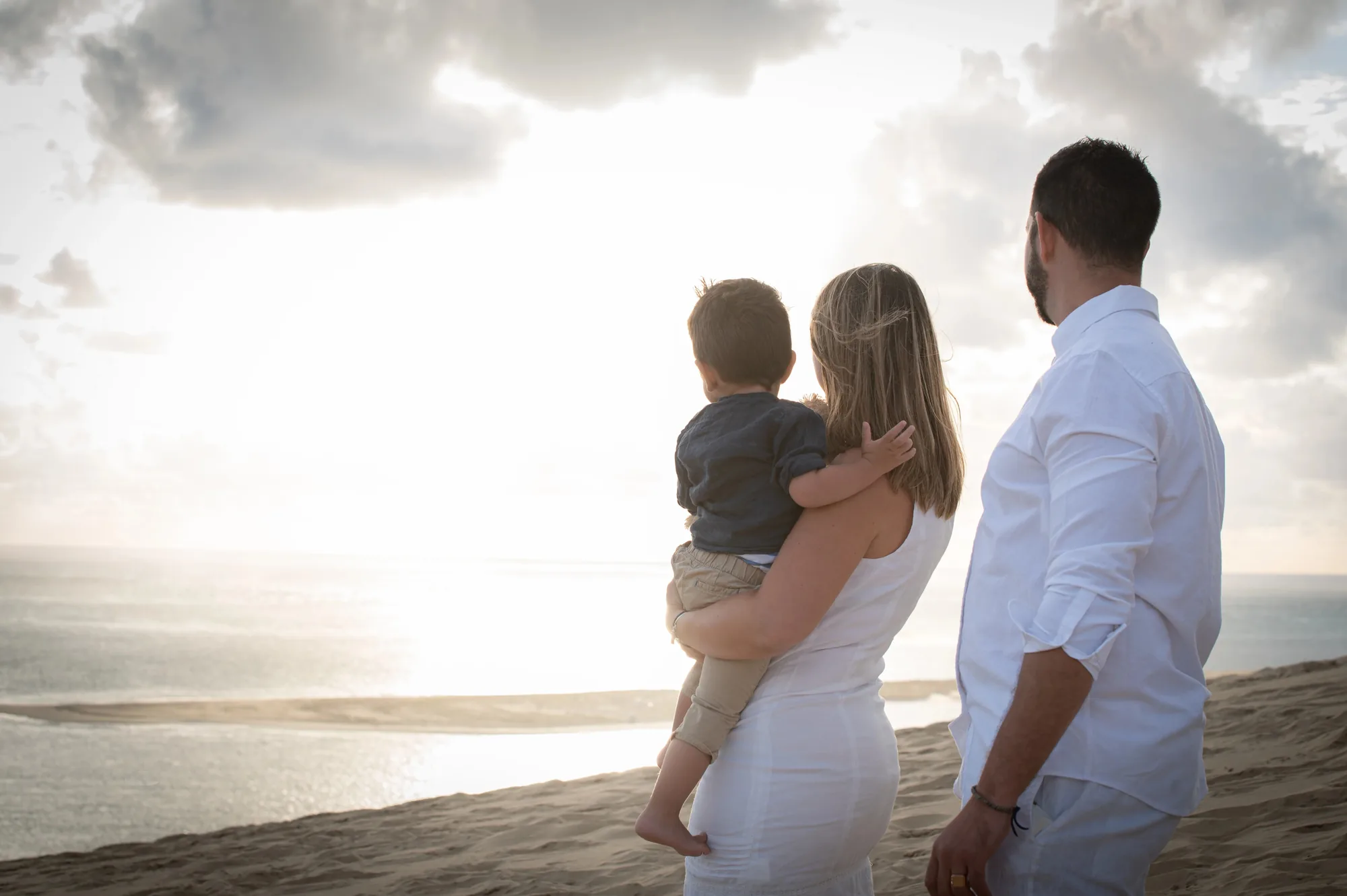 Famille de dos face à l'océan, mère portant son enfant, coucher de soleil