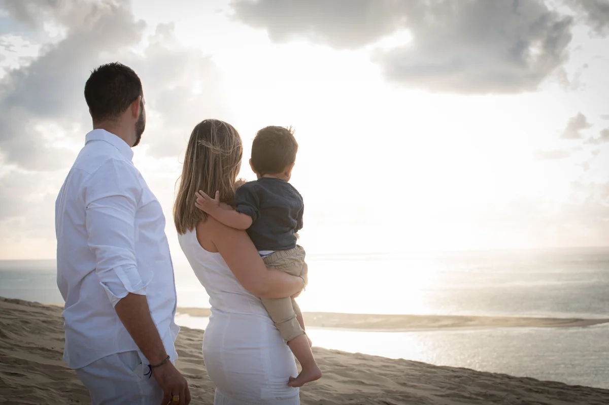 Famille de trois personnes de dos face à l'océan au coucher du soleil, père tenant son enfant dans les bras