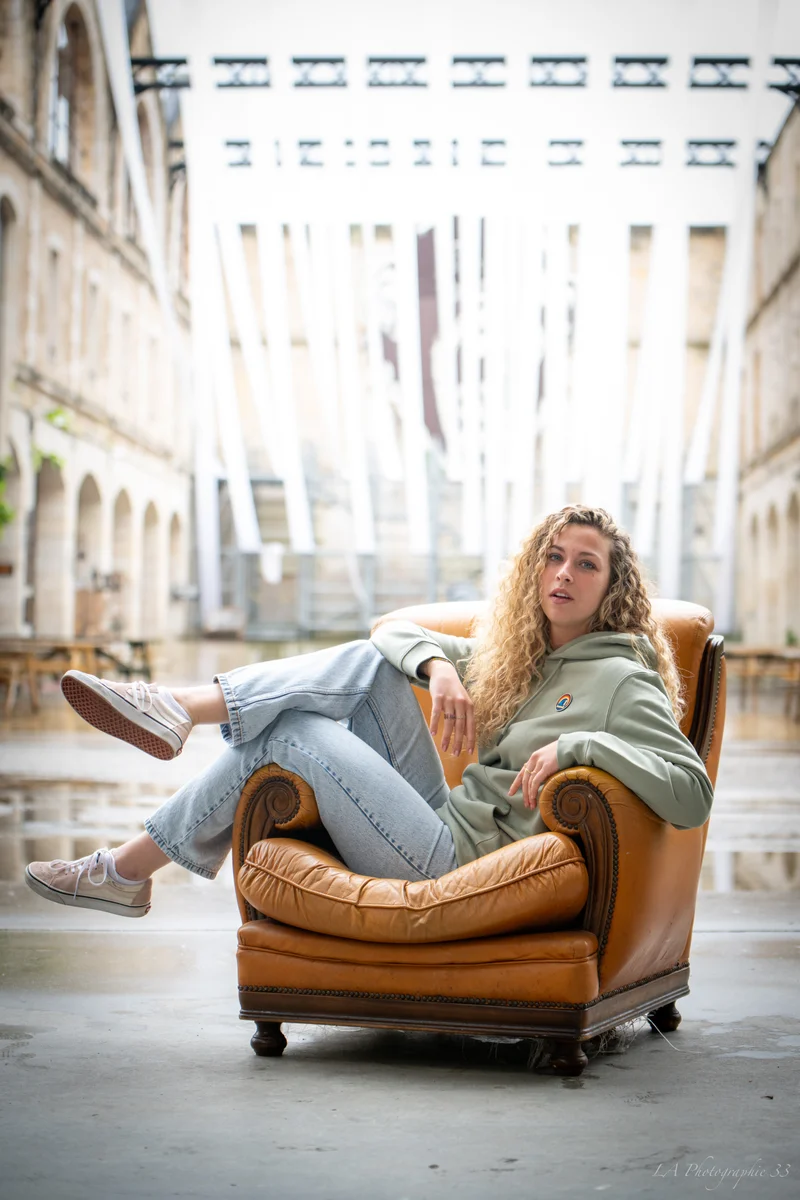Jeune femme aux cheveux bouclés assise dans un fauteuil en cuir vintage, jambe repliée, cour intérieure en pierre